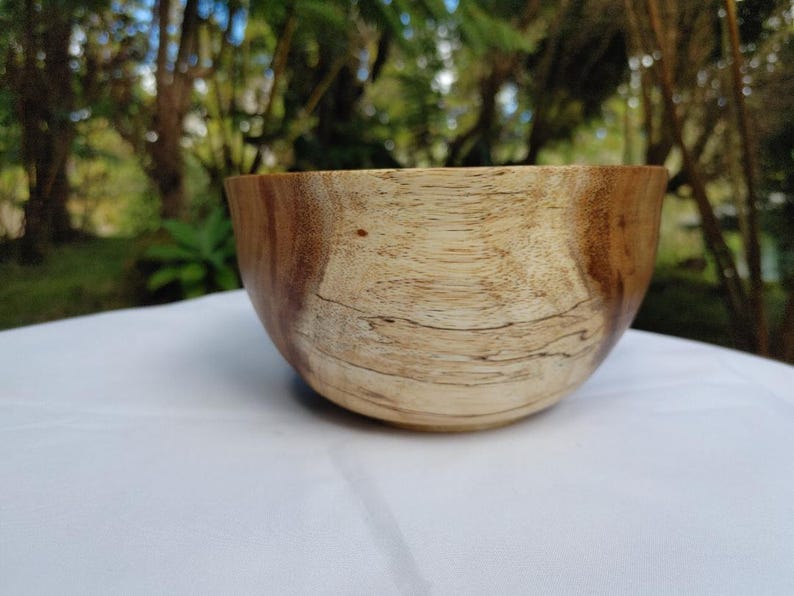 May include: A wooden bowl with a natural finish. The bowl has a light brown interior and a darker brown exterior. The wood grain is visible. The bowl is sitting on a white surface.