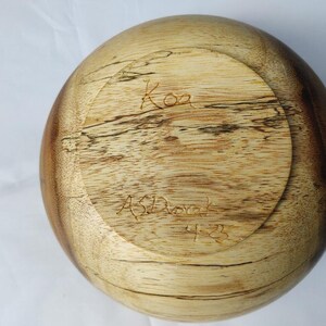 May include: A wooden bowl with a rounded shape, featuring a light brown and dark brown wood grain pattern. The bottom of the bowl has the word "Koa" and the name "AS Dorak" carved into the wood, along with the number "4-23".