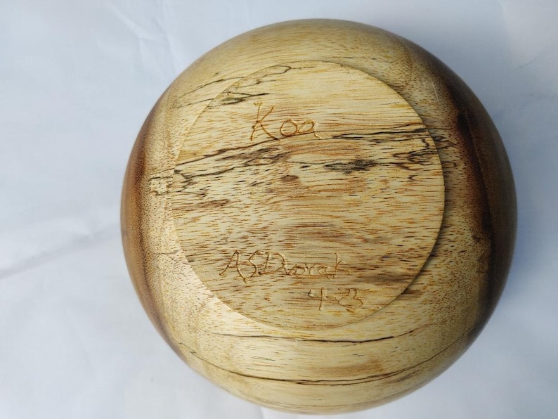 May include: A wooden bowl with a rounded shape, featuring a light brown and dark brown wood grain pattern. The bottom of the bowl has the word "Koa" and the name "AS Dorak" carved into the wood, along with the number "4-23".