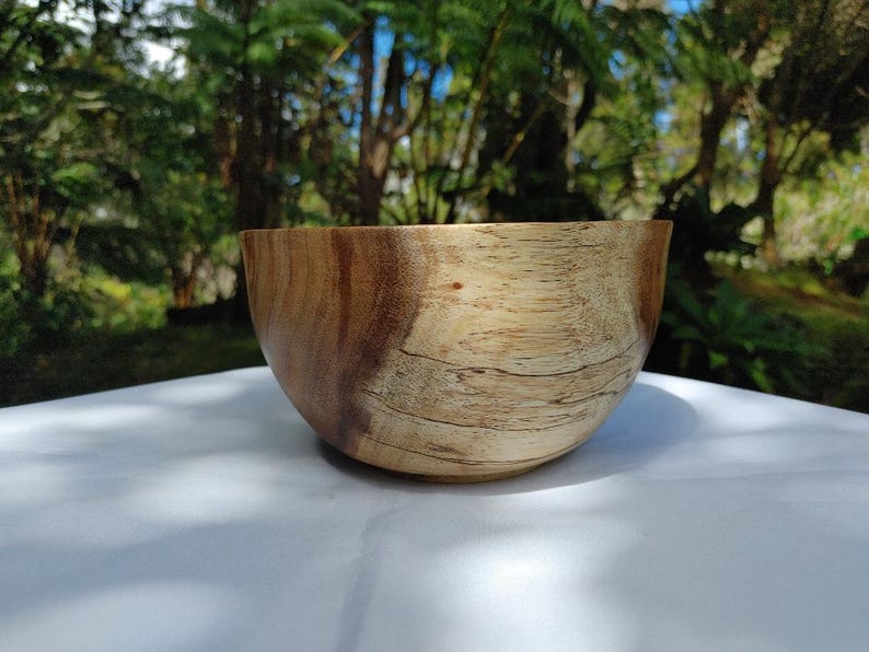 May include: A wooden bowl with a natural wood grain pattern. The bowl is light brown with darker brown streaks and a smooth, curved shape. It sits on a white surface, with a blurred green background.