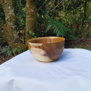 May include: A handcrafted wooden bowl with a natural finish. The bowl is made of light and dark brown wood, showcasing the grain. It sits on a white surface, with a blurred green background of trees and foliage.