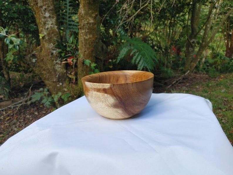 May include: A handcrafted wooden bowl with a natural finish. The bowl is made of light and dark brown wood, showcasing the grain. It sits on a white surface, with a blurred green background of trees and foliage.
