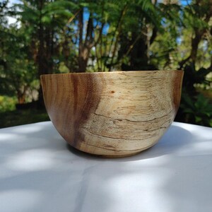 May include: A handcrafted wooden bowl with a natural finish. The bowl has a rounded shape and a light brown color with darker wood grain patterns. The bowl is sitting on a white surface, with a blurred green background.