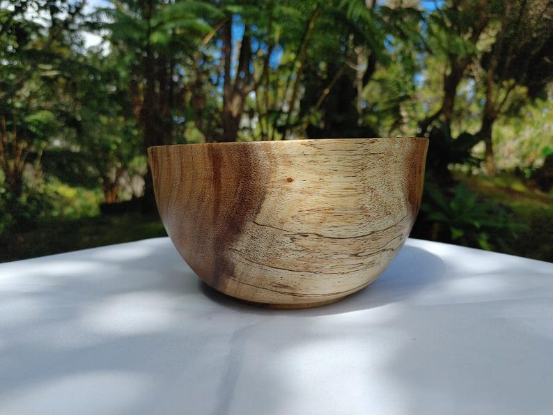 May include: A handcrafted wooden bowl with a natural finish. The bowl has a rounded shape and a light brown color with darker wood grain patterns. The bowl is sitting on a white surface, with a blurred green background.