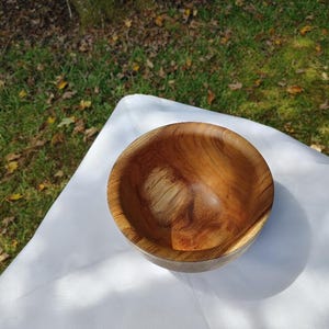 May include: A handcrafted wooden bowl with a natural wood grain pattern. The bowl is round with a smooth interior and a slightly rougher exterior. The bowl is set on a white cloth, with a grassy background.