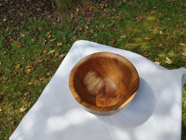 May include: A handcrafted wooden bowl with a natural wood grain pattern. The bowl is round with a smooth interior and a slightly rougher exterior. The bowl is set on a white cloth, with a grassy background.