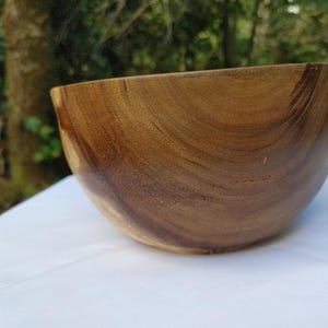 May include: A wooden bowl with a smooth, curved design. The bowl is made of a warm, brown wood with visible grain patterns. The bowl is set against a white surface with a blurred green background.