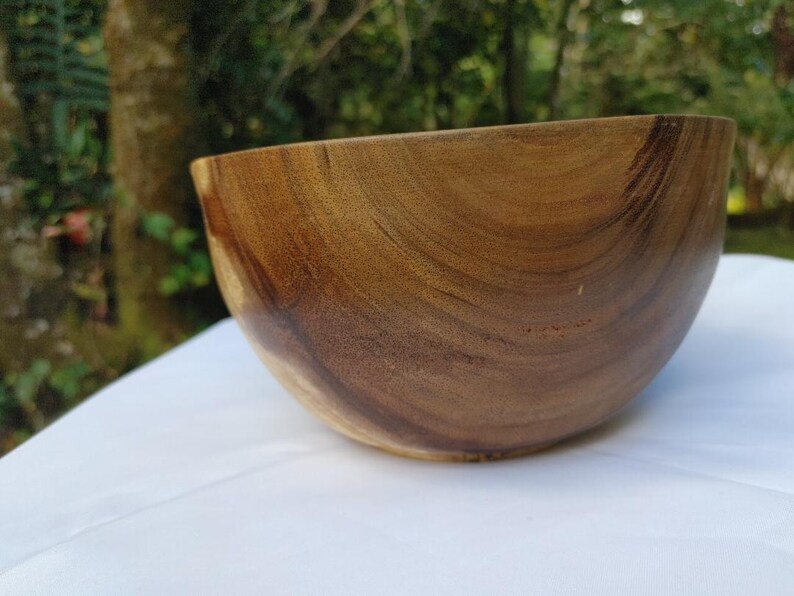 May include: A wooden bowl with a smooth, curved design. The bowl is made of a warm, brown wood with visible grain patterns. The bowl is set against a white surface with a blurred green background.