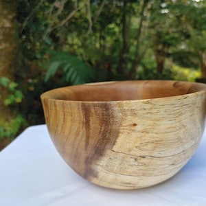May include: A handcrafted wooden bowl with a natural, light brown and dark brown grain pattern. The bowl has a smooth, rounded shape and is likely used for serving food or as a decorative item. The background is a blurred outdoor scene.