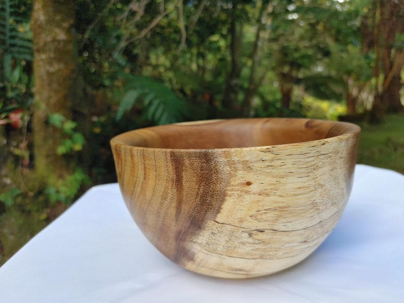 May include: A handcrafted wooden bowl with a natural, light brown and dark brown grain pattern. The bowl has a smooth, rounded shape and is likely used for serving food or as a decorative item. The background is a blurred outdoor scene.