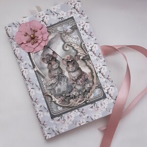 May include: A small, gray journal with a floral design and a pink flower embellishment. The journal features a vintage illustration of two mice wearing hats and dresses. A pink ribbon is attached to the journal.