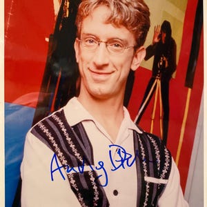 May include: A portrait of a smiling person wearing glasses and a light-colored shirt with a patterned vest. The image has an autograph in blue ink. The background features abstract shapes and a silhouette of a person with a camera.