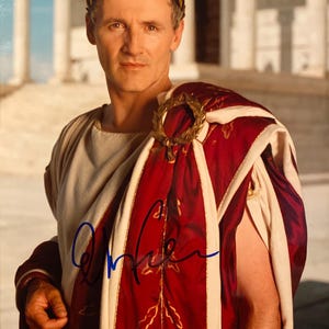 May include: A man in a Roman-style costume, wearing a laurel wreath and a red and white toga with gold accents. The image is signed in blue ink. The background features a classical building.