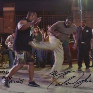 May include: A promotional photograph for the film Blood and Bone featuring a fight scene with two individuals in motion, one wearing a black tank top and patterned shorts, the other in a tan t-shirt and trousers, with a signature in black ink.