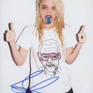 May include: A white t-shirt with a black and pink graphic of a man wearing glasses. The person in the image has blue tongue and is giving a thumbs up. The image is signed in blue ink.