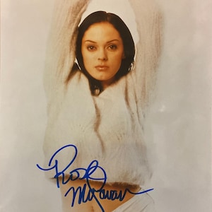 May include: A portrait of a person wearing a white, fuzzy sweater with arms raised. The person has dark hair and is looking at the camera. A blue signature is visible on the lower part of the image.