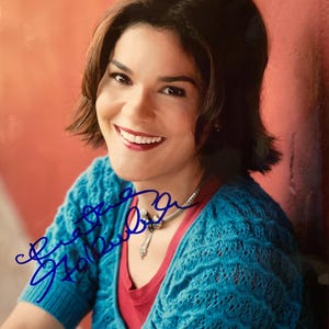 May include: A person with dark hair smiles, wearing a blue knitted cardigan over a red top. A silver necklace with a pendant is visible. The image includes a blue signature. The background is a warm orange-red.