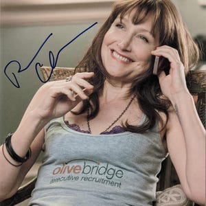 May include: A woman wearing a gray vest top with the text "olive bridge executive recruitment" and a signature. She is smiling and holding a mobile phone to her ear. She has dark hair and is wearing a necklace and bracelets.