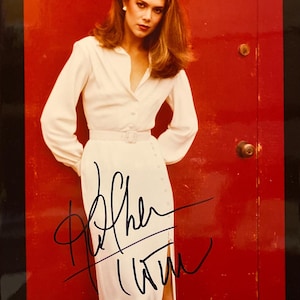 Puede incluir: Una mujer con un vestido blanco abotonado con cinturón posa frente a una puerta roja. Tiene el pelo largo y castaño y lleva un pendiente de perla. La foto está firmada con "Cher" en tinta negra.