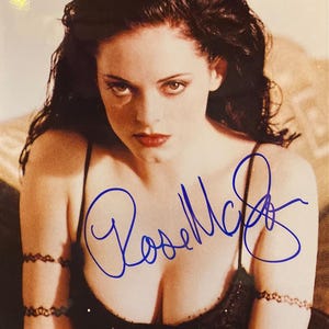 May include: A portrait of a woman with dark hair and red lipstick. She is wearing a black top and has a signature in blue ink. The image is a close-up shot, focusing on her face and upper body.