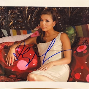 Puede incluir: Fotografía autografiada de una mujer con un vestido color crema, sentada en una cama con cojines decorativos. Los cojines tienen diseños rojos y marrones. La foto está firmada con tinta azul.