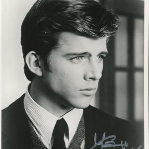 May include: A black and white portrait photograph of a person with dark, styled hair, wearing a collared shirt with a striped pattern, a dark necktie, and a textured sweater vest under a dark blazer, featuring a handwritten signature in the corner.