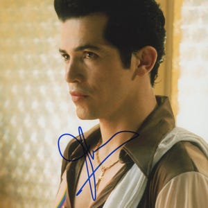 May include: A glossy photograph featuring a person with dark hair styled in a pompadour, wearing a brown collared shirt with a white cloth draped over the shoulder and a gold chain necklace. A blue ink signature is written across the chest area.