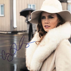 May include: A woman wearing a cream-coloured hat and coat with a white fur collar. The image has a signature in the lower left corner. The background is blurred, showing a person holding an umbrella and buildings.