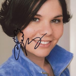 May include: A person with dark hair and brown eyes smiles, wearing a blue zip-up jacket with a high collar and a zipper. The image includes a signature in black ink.