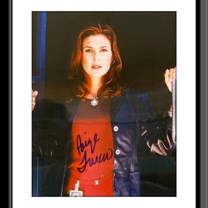 Op de afbeelding: Een gesigneerde foto van een vrouw met lang bruin haar, die een rood shirt en een zwart leren jack draagt. De vrouw kijkt recht in de camera. De handtekening is in paarse inkt en luidt "Paige Turco".