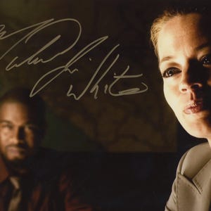 May include: A glossy promotional photograph featuring a close-up of a light-skinned individual in a beige blazer and a blurred dark-skinned individual in a dark shirt in the background. The image includes a prominent silver ink autograph reading Michael Jai White.
