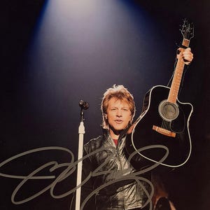 Könnte beinhalten: Ein Mann hält eine schwarze Akustikgitarre, beleuchtet von einem Scheinwerfer. Er trägt eine schwarze Lederjacke und ein rotes Hemd. Das Bild enthält ein Autogramm.