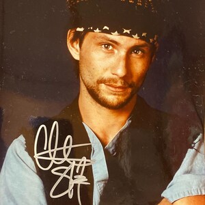 Könnte beinhalten: Porträt eines Mannes mit schwarzer Weste über einem hellblauen Hemd und einem gemusterten Hut. Das Bild enthält ein Autogramm in weißer Tinte. Der Mann hat einen Bart und schaut direkt in die Kamera.