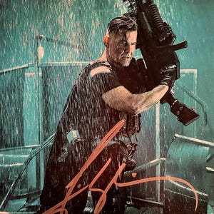 May include: A man in a black shirt and tactical gear holds a large weapon in the rain. The image has a teal and black color scheme, with water droplets visible. An orange signature is in the foreground.