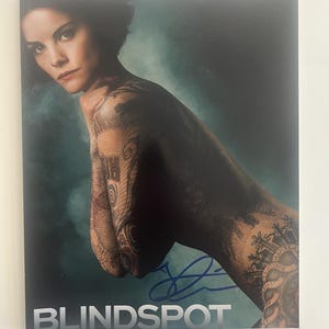 Puede incluir: Una impresión promocional para la serie de televisión Blindspot. La impresión presenta a una mujer con intrincados tatuajes que cubren sus brazos y torso. El título del programa está impreso en letras blancas en la parte inferior, y hay una firma azul.