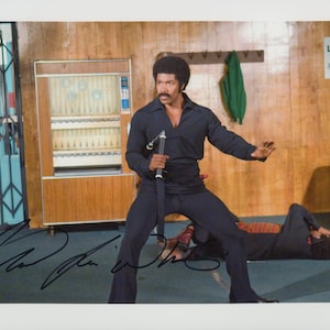 May include: A person with an afro hairstyle wearing a black long-sleeved shirt and matching trousers, holding nunchucks in a defensive stance. A second person lies on the floor in the background. A black signature is written across the bottom.