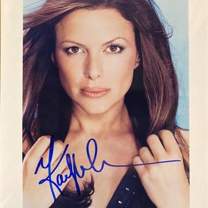 May include: A portrait photograph of a woman with long brown hair, looking directly at the camera. The image is signed in blue ink. The woman is wearing a dark top. The background is a light blue.