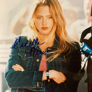 May include: A photograph of a woman with long blonde hair wearing a denim jacket over a pink top. She is wearing a watch with a black strap. The image is signed in blue ink with the name "Estella Warren".