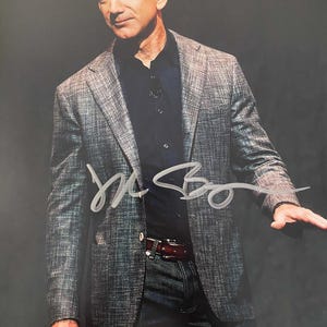 Op de afbeelding: Gesigneerde foto van een man in een grijze blazer over een donkerblauw overhemd en een blauwe jeans. De handtekening is in zilverkleurige inkt. De man is kaal en heeft een serieuze uitdrukking. Een bruine leren riem met een zilveren gesp is zichtbaar.