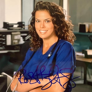 May include: A woman wearing a blue medical scrub top with a stethoscope around her neck. Her arms are crossed, and she is smiling. The image has an autograph in blue ink across the front.