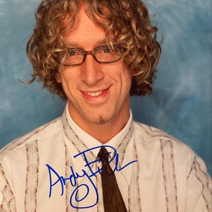 May include: A smiling person with curly light brown hair and glasses. They are wearing a white collared shirt with vertical stripes and a dark brown tie. The image includes a blue signature that reads "Andy Fickman".