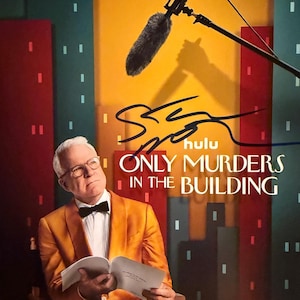 Puede incluir: Póster de "Only Murders in the Building" con Steve Martin. Viste una chaqueta naranja y pajarita, sosteniendo un libro. El póster incluye el logo de Hulu y el texto: "New Season Aug 27". Se ve un micrófono.