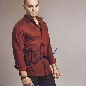 Puede incluir: Un hombre con una camisa roja oscura con mangas enrolladas, pantalones negros y un reloj dorado. Una firma es visible en la camisa. El fondo es gris liso.