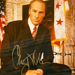 May include: A signed photograph of a man in a dark pinstripe suit, white shirt, and red tie. The signature is in gold ink. The background includes an American flag and a flag with red stars.