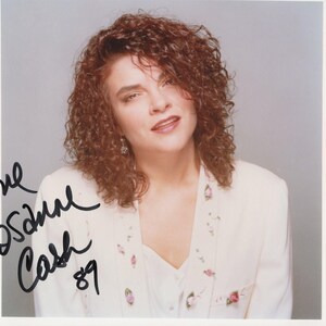 May include: A portrait of Rosanne Cash, signed with the inscription "Love Rosanne Cash '89". She is wearing a white jacket with floral embroidery. Her curly auburn hair frames her face, and she is smiling.