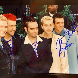 May include: A group of six men posing for a photo, likely a boy band. The men are wearing various outfits, including leather jackets, suits, and patterned shirts. The photo has several signatures in blue ink.