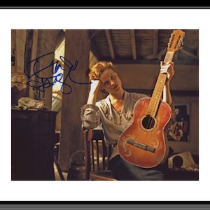 May include: A framed photograph of a woman holding a vintage acoustic guitar. The guitar has a reddish-brown finish with decorative carvings. The woman is seated, wearing a light-coloured blouse, and appears to be deep in thought. The image is signed in blue ink.