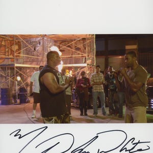May include: A photograph featuring two individuals facing each other in an outdoor setting with scaffolding and a fire effect in the background, accompanied by a black ink signature at the bottom of the white border, signed Michael Jai White.
