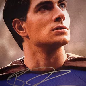 May include: A portrait of a man in a Superman costume, with a blue shirt and a brown cape. The image includes a signature in silver ink. The man is looking up and to the right.