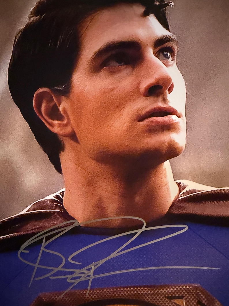 May include: A portrait of a man in a Superman costume, with a blue shirt and a brown cape. The image includes a signature in silver ink. The man is looking up and to the right.